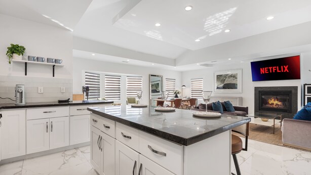Een moderne keuken met witte kastjes, een marmeren aanrechtblad en een inbouw eiland. Er is een zithoek met een tafel en stoelen, en een woonkamer met een bank en een open haard.
