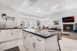 A modern kitchen with white cabinetry, a marble countertop, and a built-in island. There is a dining area with a table and chairs, and a living room with a sofa and a fireplace. - Minimalist Serene Home in near Rose Bowl Stadium  Pasadena (Pasadena)