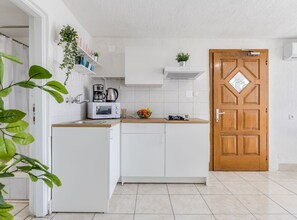 Eine kleine Küche mit weißen Schränken, einer Holztür und einer Arbeitsplatte mit Wasserkocher und Obstschale.
