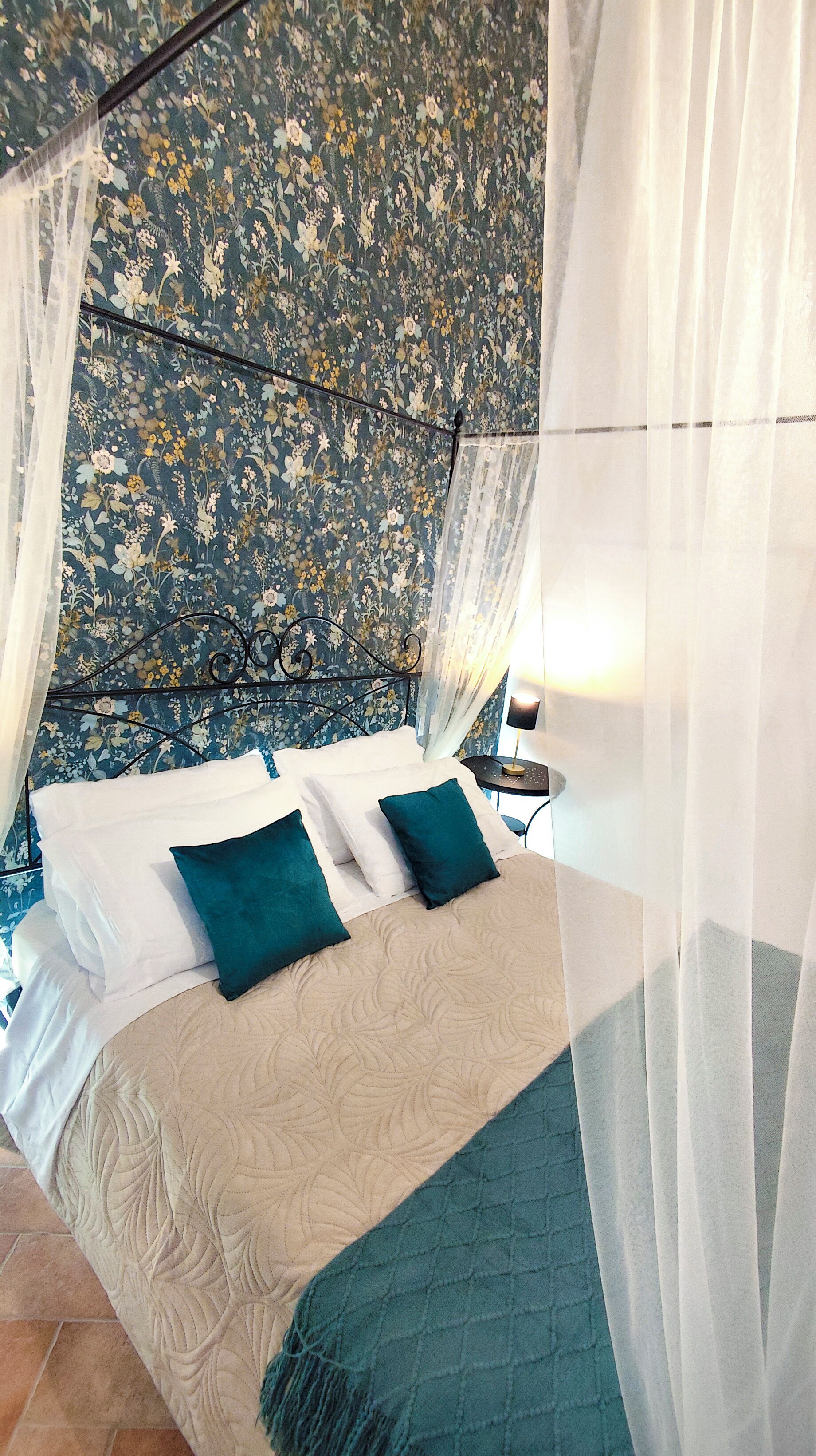 A bed with a floral headboard, a bedside table with a lamp, and sheer curtains.