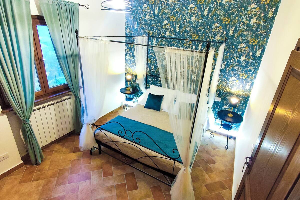 A bedroom with a four-poster bed, a radiator, a small table with a lamp, and a floral wallpaper.