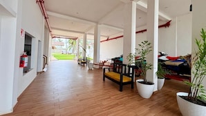 Um corredor amplo com piso de madeira, bancos, plantas em vasos e um extintor de incêndio na parede.