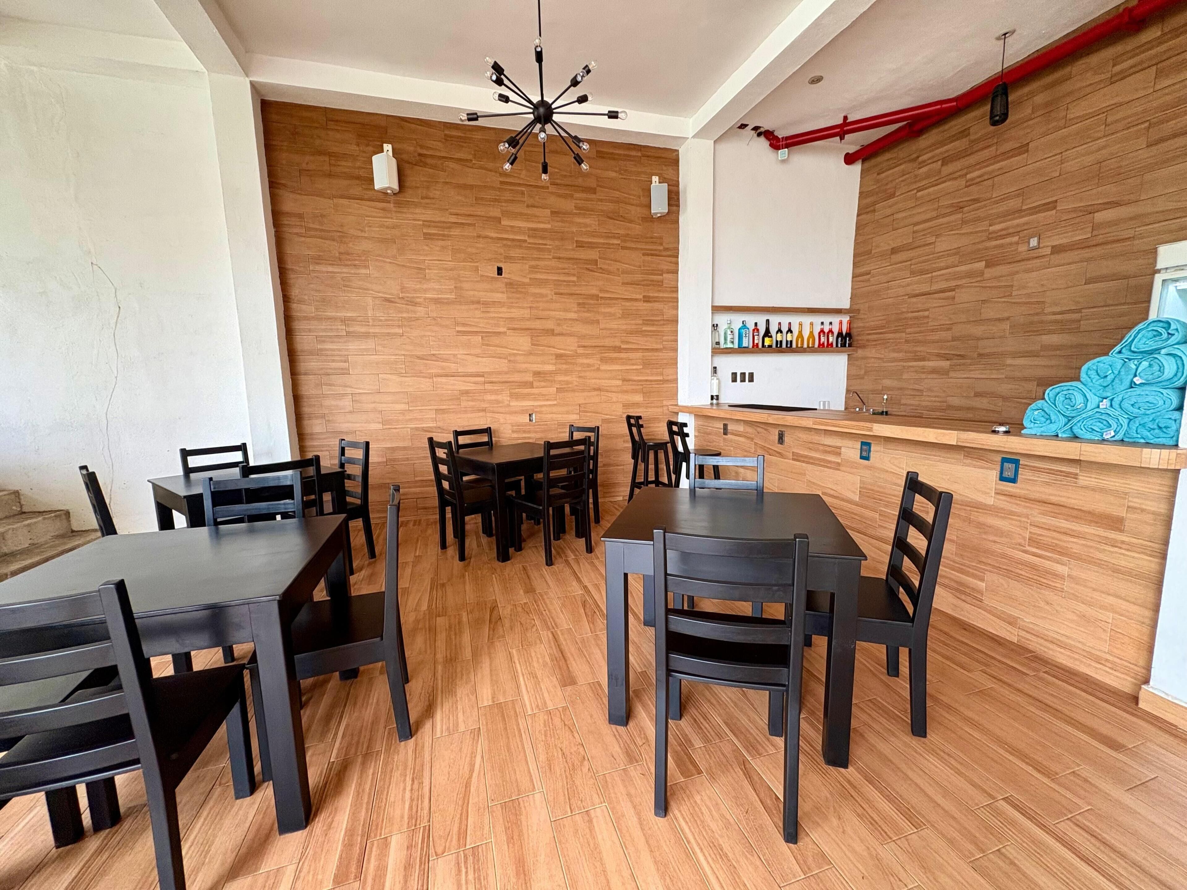 Een eetgedeelte met houten vloer, zwarte tafels en stoelen, een centrale kroonluchter, een bar met flessen en een houten wand.