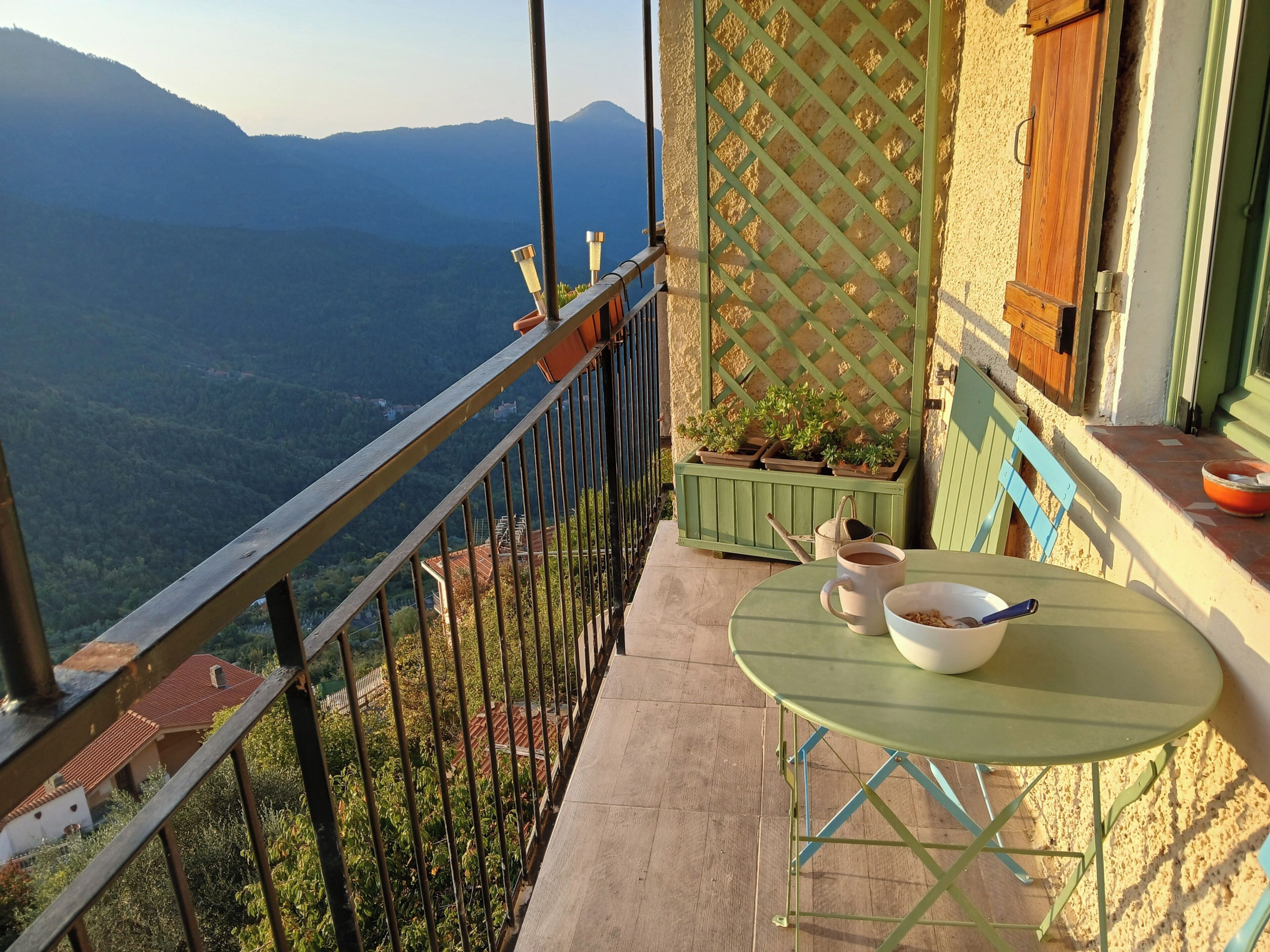A balcony with a view of a mountainous landscape, a table with a bowl and a cup, and a green lattice partition.