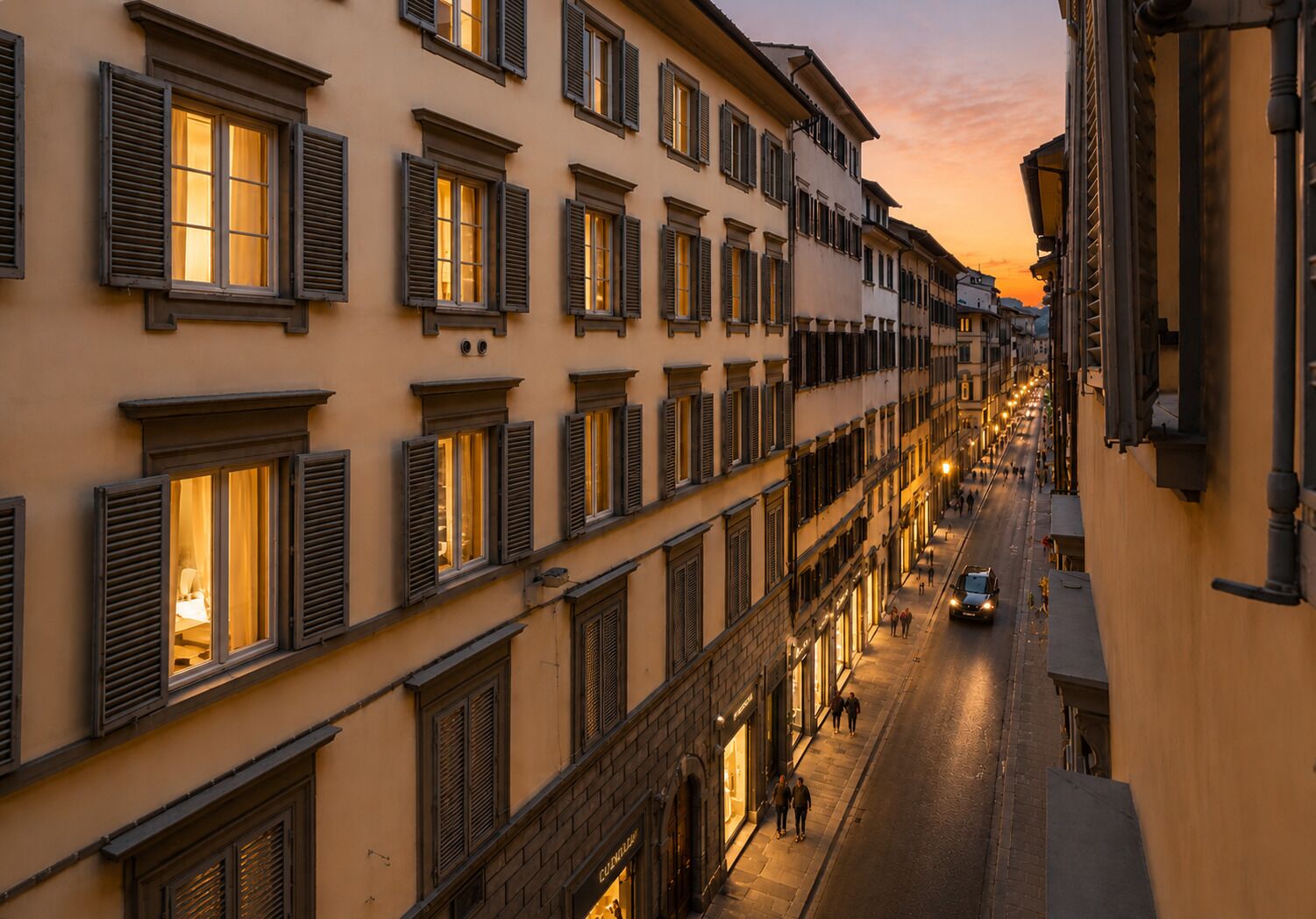 A street view of a building with multiple windows, some with shutters, and a few storefronts with illuminated signs.