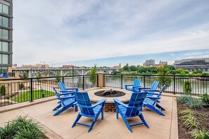 Une terrasse dotée de chaises Adirondack bleues et d’un foyer, offrant une vue sur une rivière et le paysage urbain.