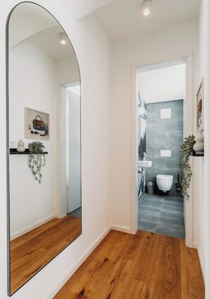 A hallway with a wooden floor, a large mirror, and a door leading to a bathroom with a sink and toilet. - Castle on Lake Constance I Private Sauna & Hot Tub (Stockach)