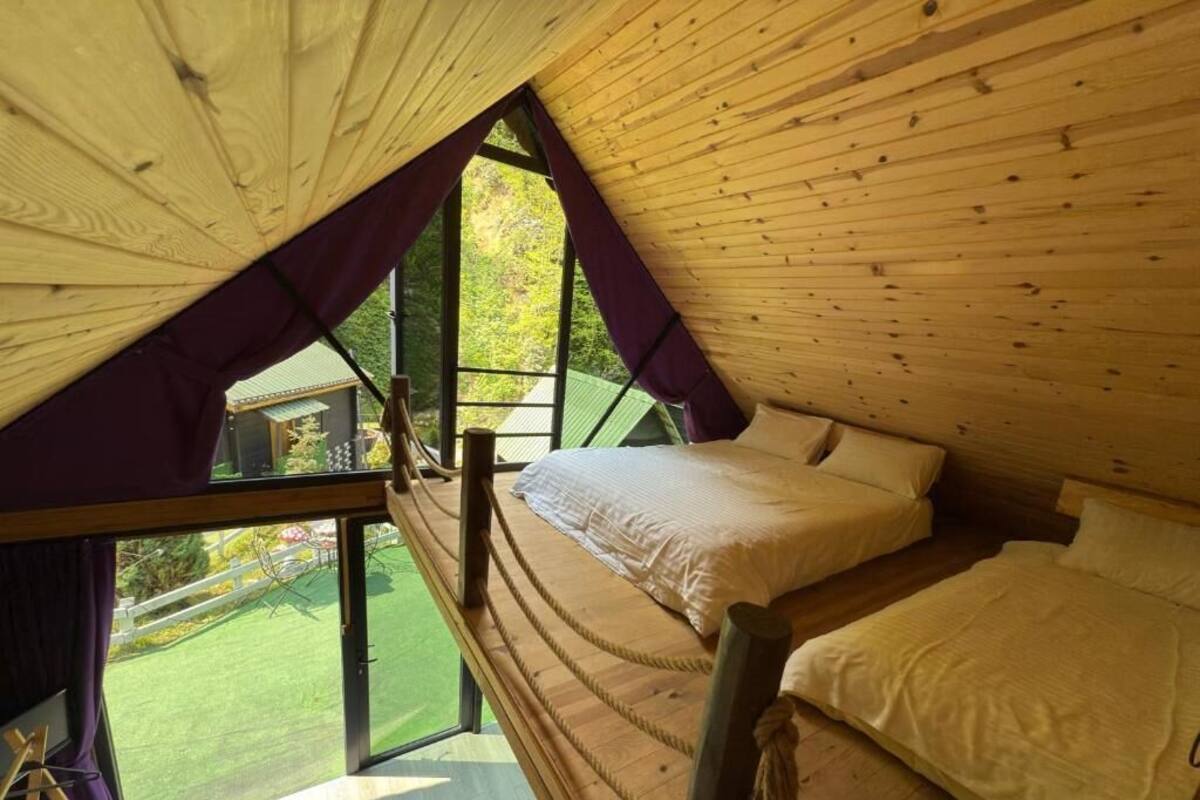 A bedroom with a wooden sloped ceiling, a bed with white bedding, and a large window offering a view of a green landscape.