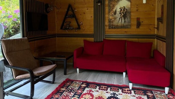 A wooden interior with a red sofa, a chair, a table, and a patterned rug. - Ayder Dağ Evleri Elizan (Camlihemsin)