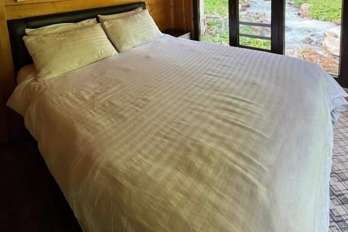 A wooden cabin bedroom with a bed, a wall-mounted light, and a window with a view of greenery.