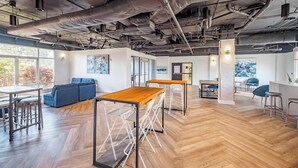 Ein geräumiges Büro mit hohen Decken, freiliegenden Leitungen und verschiedenen Sitzbereichen, darunter ein blaues Sofa und ein Holztisch mit Hocker.