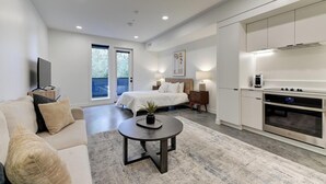 Quarto de hotel moderno com uma cama, área de estar com sofá e mesa de centro, cozinha com fogão e forno, e grandes janelas.