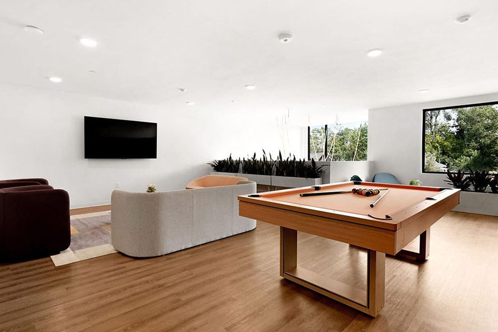 A modern living room with a pool table, comfortable seating, a flat-screen TV, and large windows offering a view of greenery.