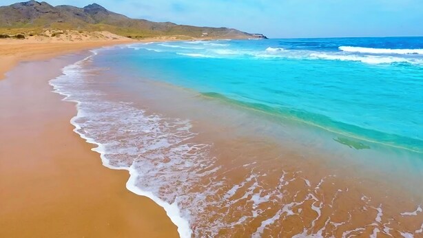 Uma praia com águas cristalinas, areal arenoso e montanhas ao longe.