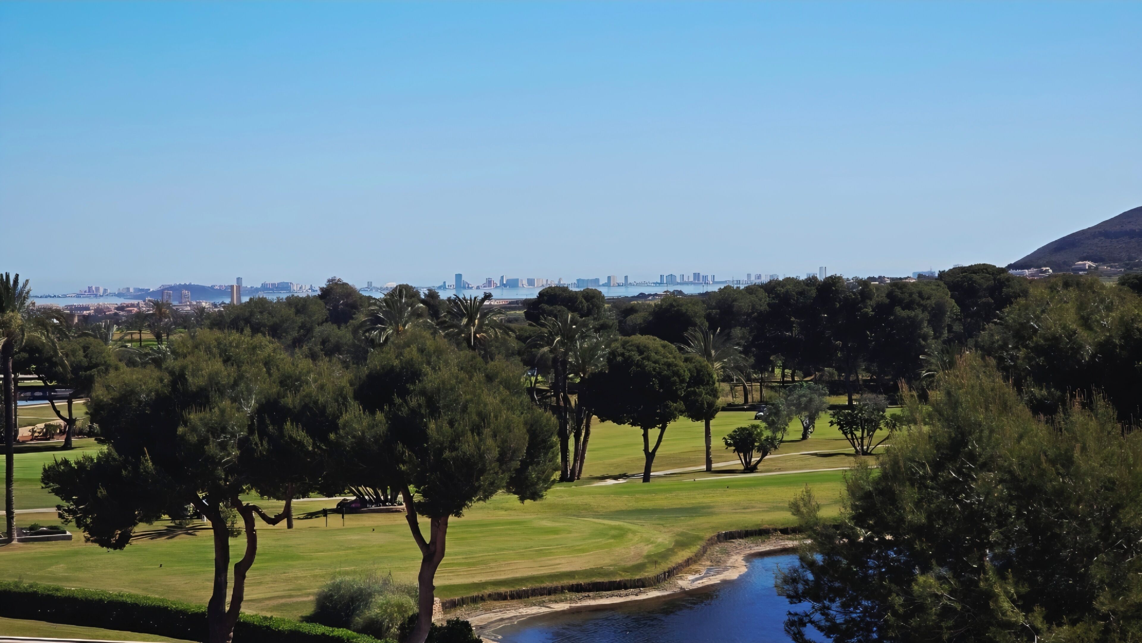 Ein Golfplatz mit einem Wasserteil, Bäumen und einer entfernten Skyline der Stadt.