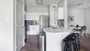 A modern kitchen with white cabinetry, a stainless steel refrigerator, a built-in island with bar stools, and a dining area with a table and chairs. - Landing | Stylish 2BD, Gym, Tennis Court (Stockbridge)