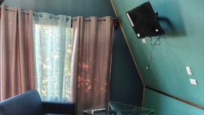 Ett rum med en blå soffa, en vit stol, ett glasbord, en väggmonterad tv och ett fönster med gardiner.