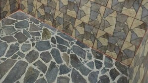 Ett badrum med en vägg och ett golv som är beklätt med ett stenmosaikmönster, ett duschmunstycke i metall och ett handfat i ett hörn.