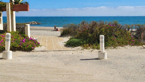 Ein Küstenweg führt zu einem Strand mit freiem Blick auf den Ozean. In der Ferne sind einige Personen zu sehen, und ein Gebäude mit rotem Dach.
