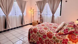 Une chambre à coucher avec un lit, des rideaux, une table de chevet, une lampe et une fenêtre.