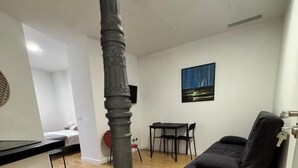 A room with a wooden floor, a black sofa, a dining table with chairs, a painting on the wall, and a pillar. - Apartamentos Melisa (Madrid)