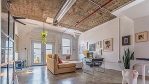 A spacious living area with a large sofa, a desk, and a chair. There are framed pictures on the walls and a hanging plant. - Bright and airy loft space 10 min walk from stadiums with private roof terrace (Atlanta)