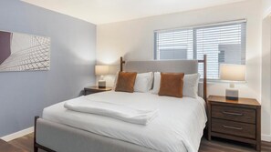 A neatly made bed with white bedding and brown pillows, two bedside tables with lamps, a window with blinds, and a piece of abstract art on the wall. - Landing | Amazing 1BD in Glendale (Glendale)