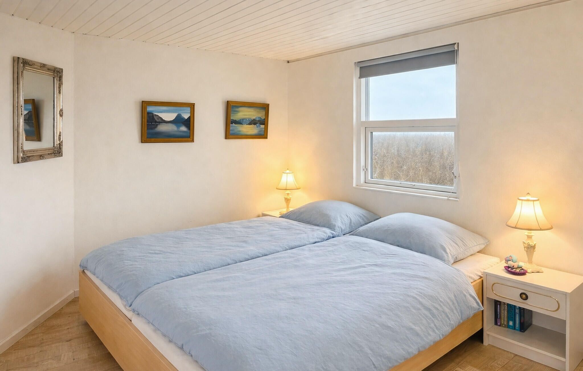 A bedroom with a double bed, two bedside lamps, a nightstand with books, and two framed paintings on the wall.