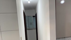 A narrow hallway with white tiled flooring, a black bench on the left, and a black ottoman on the right. - Cozy apartment (Salvador)