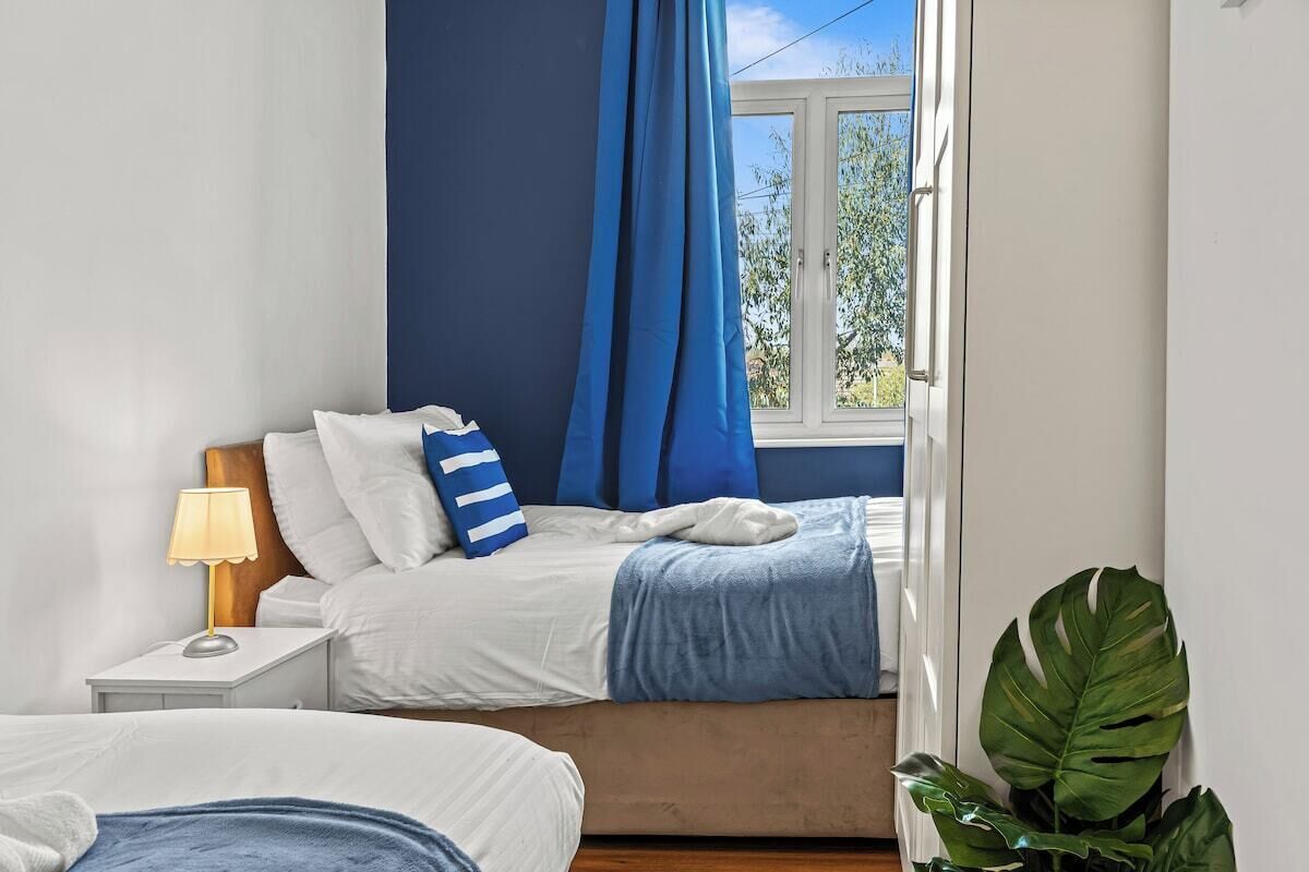 A bedroom with a bed, a nightstand, a lamp, a window with blue curtains, and a plant.