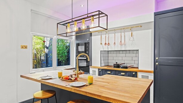 Une cuisine moderne avec un îlot central en bois, des tabourets de bar, un luminaire suspendu et une vue sur la verdure à l’extérieur.