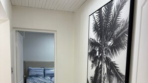 Um corredor com uma cama ao fundo, um quadro grande emoldurado com palmeiras e uma porta à direita.