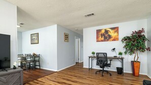 Una habitación amplia con piso de madera, un comedor con sillas, un escritorio con silla, un televisor y una planta en maceta.