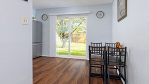 Una habitación con piso de madera, una mesa de comedor con sillas, un patio trasero con puerta corrediza de vidrio y un refrigerador pequeño.