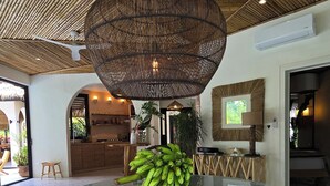 Quarto amplo com uma mesa de vidro redonda, luminária de teto em palha e uma cozinha moderna ao fundo.