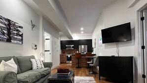 Un soggiorno moderno con divano, tavolino, televisore piatto e cucina sullo sfondo.