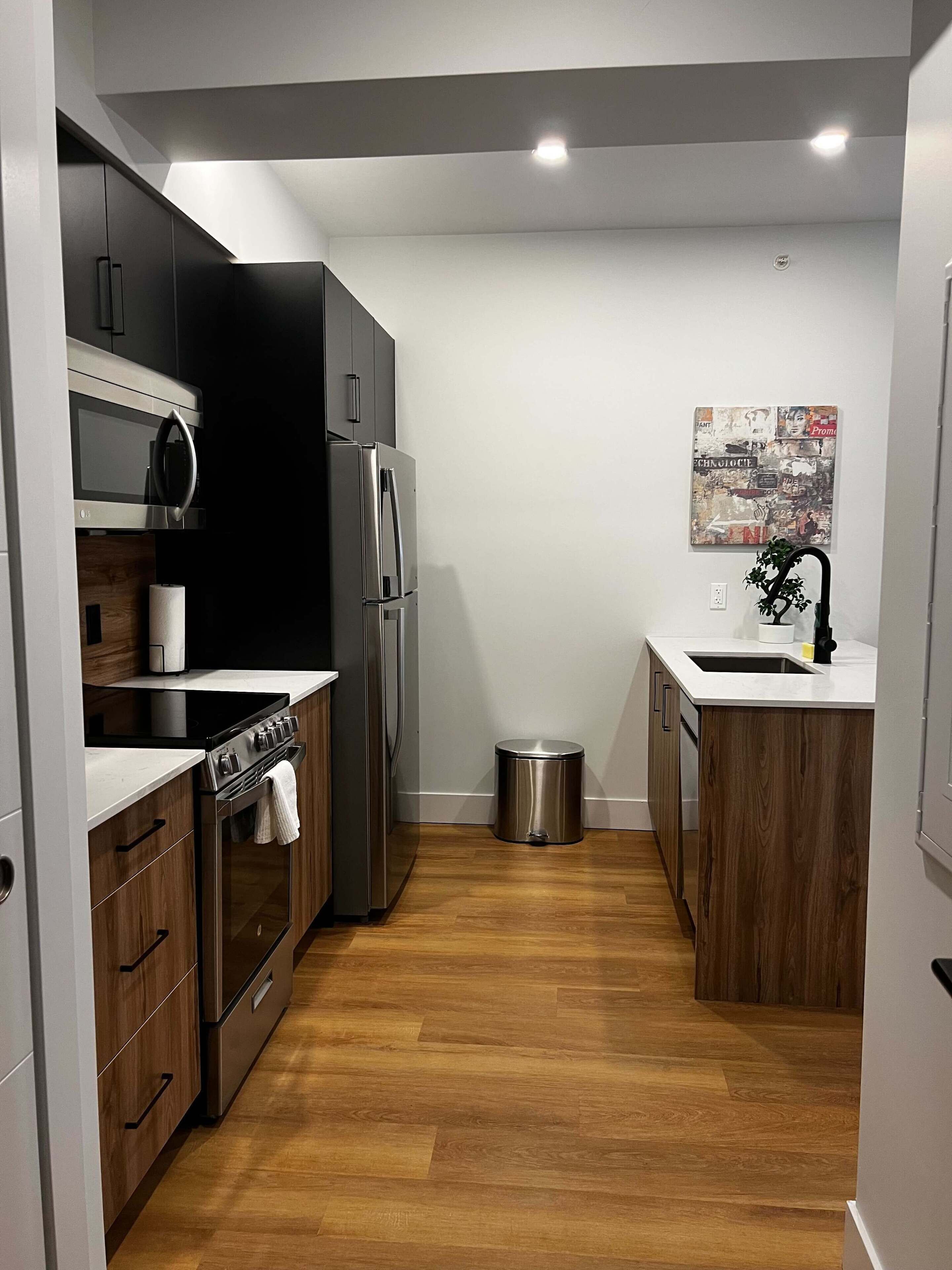 A compact kitchen with modern appliances, wooden cabinets, and a small trash bin.