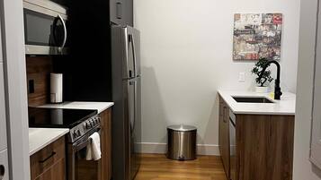 A compact kitchen with modern appliances, wooden cabinets, and a small trash bin.