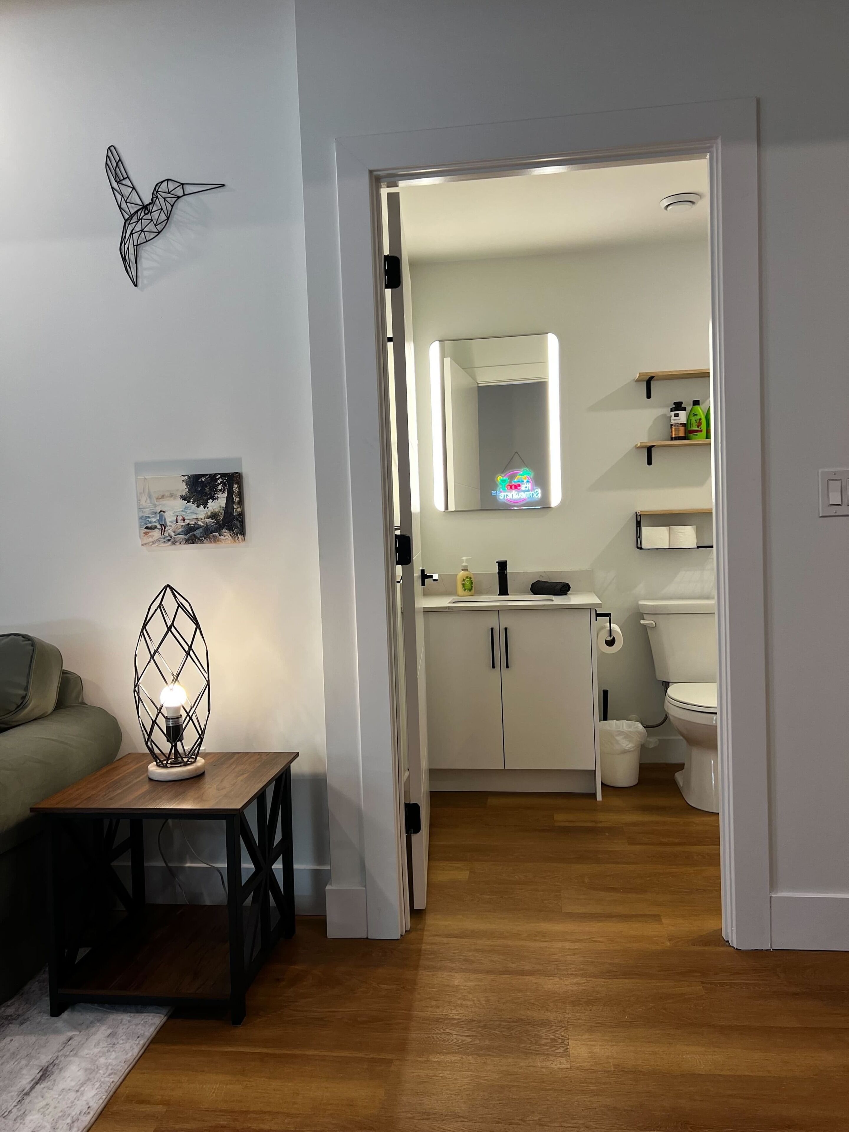 A modern bathroom with a toilet, sink, and mirror, and a hallway leading to a living area with a lamp and artwork.