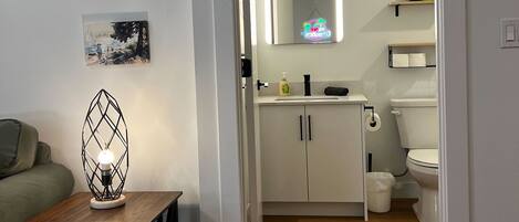 A modern bathroom with a toilet, sink, and mirror, and a hallway leading to a living area with a lamp and artwork.