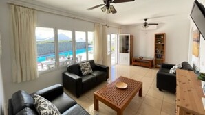 A spacious living room with a large window, a ceiling fan, a wooden coffee table, and a bookshelf. - Villa Heartly – Luxury, Tranquillity & Panoramic Sea Views (Playa Blanca)