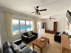 A spacious living room with a large window, a ceiling fan, a wooden coffee table, and a bookshelf. - Villa Heartly – Luxury, Tranquillity & Panoramic Sea Views (Playa Blanca)