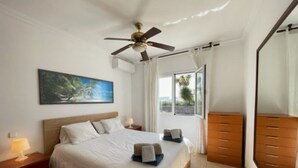 A hotel room with a bed, a nightstand, a dresser, a ceiling fan, a painting, and a window with curtains. - Villa Heartly – Luxury, Tranquillity & Panoramic Sea Views (Playa Blanca)