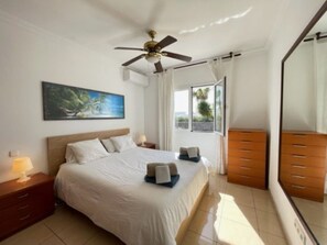 A hotel room with a bed, a nightstand, a dresser, a ceiling fan, a painting, and a window with curtains. - Villa Heartly – Luxury, Tranquillity & Panoramic Sea Views (Playa Blanca)