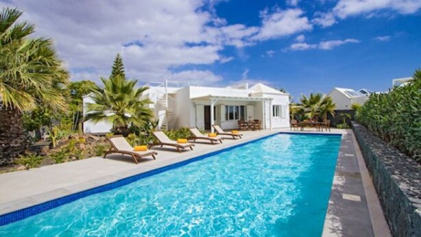 A swimming pool with lounge chairs, a white building, and tropical vegetation. - Villa Heartly – Luxury, Tranquillity & Panoramic Sea Views (Playa Blanca)