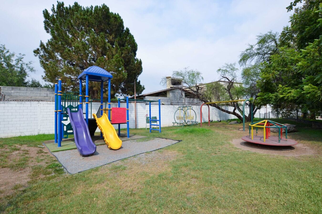 A playground with slides and swings, surrounded by a white wall and trees.