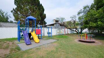 A playground with slides and swings, surrounded by a white wall and trees.