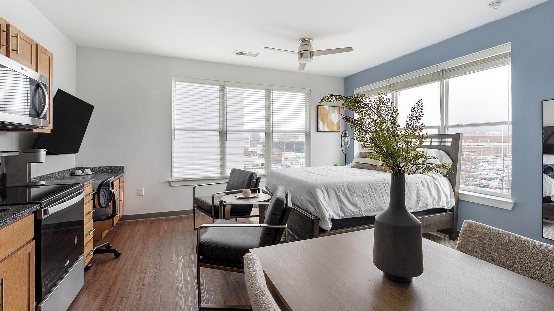 Una habitación de hotel moderna con cama, zona de comedor, kitchenette y ventanas amplias.
