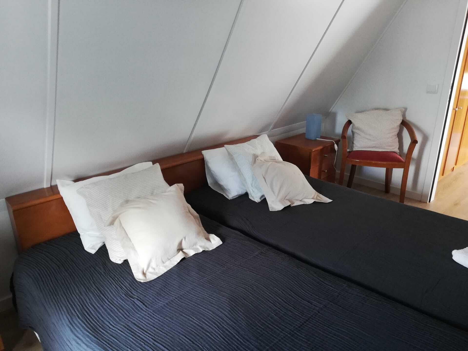 Una cama individual con colchón azul oscuro y dos almohadas, cabecera de madera, mesita de noche de madera con una lámpara, y una silla con cojín.