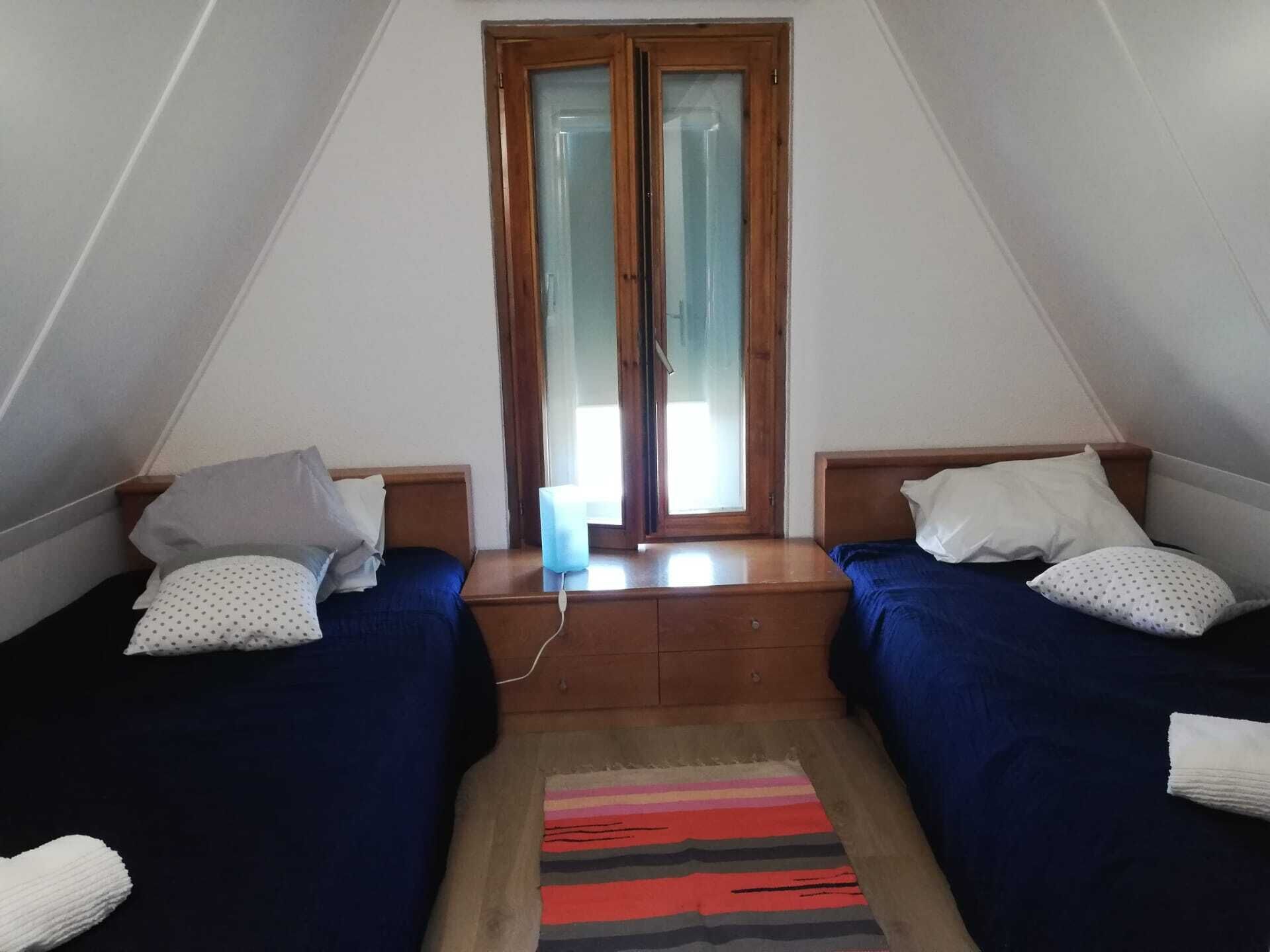 A small, triangular room with two single beds, a window with a wooden frame, and a small table between the beds.